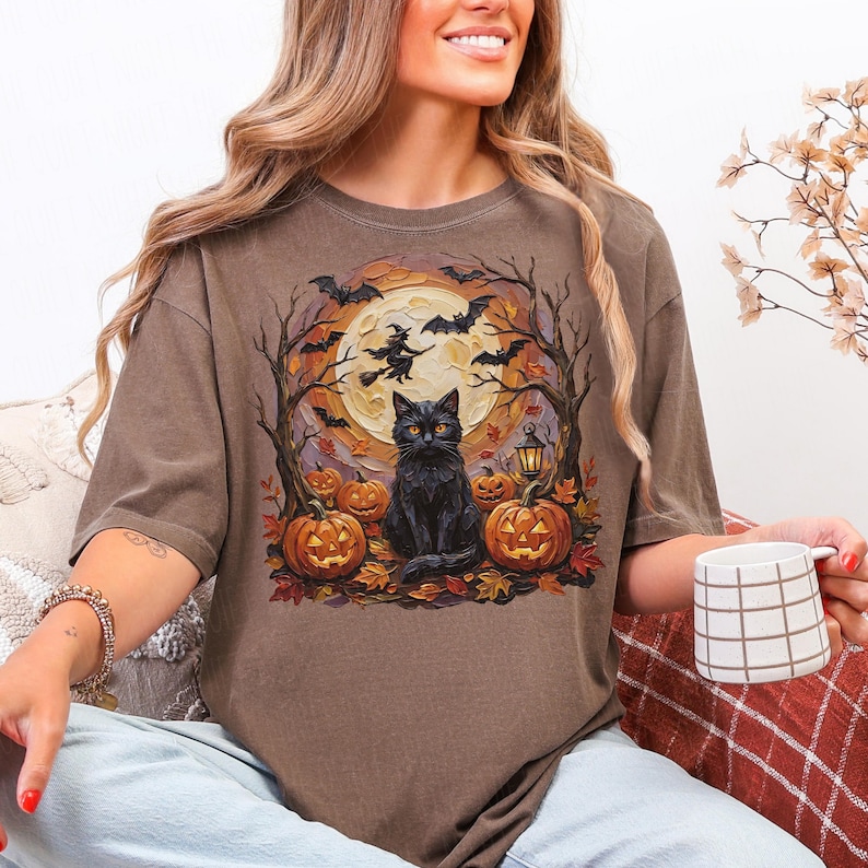 May include: A brown t-shirt featuring a graphic print of a black cat sitting in front of a large yellow moon. The design includes silhouettes of flying bats, a witch on a broomstick, bare trees, a lantern, and several orange jack-o'-lanterns.
