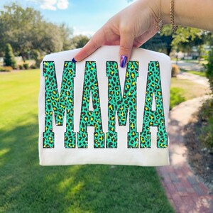 K&ouml;nnte beinhalten: Ein wei&szlig;es T-Shirt mit dem Wort MAMA in gro&szlig;en Blockbuchstaben. Die Buchstaben haben ein lebendiges Leopardenmuster in T&uuml;rkis und Neongelb mit einer abgenutzten, verwitterten Textur im gesamten grafischen Design.