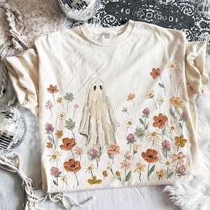 Może przedstawiać: Kremowy t-shirt z kr&oacute;tkim rękawem z centralną grafiką ducha otoczonego polem kwiat&oacute;w w stylu malarskim w odcieniach pomarańczu, ż&oacute;łci, r&oacute;żu i zieleni. Wz&oacute;r pokrywa prz&oacute;d koszulki i charakteryzuje się wyraźną teksturą nadruku.
