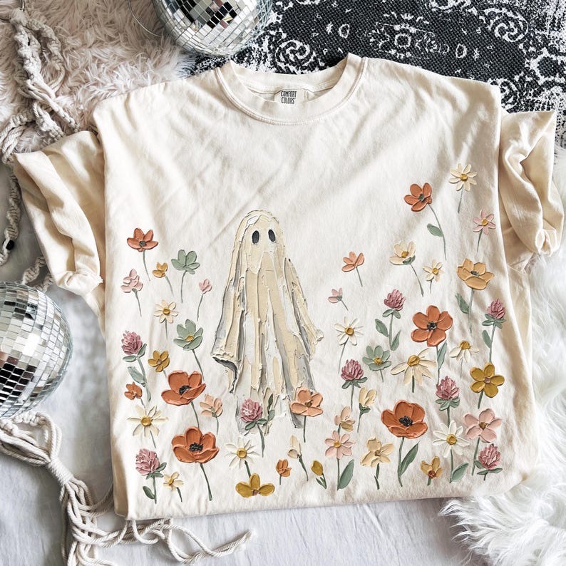 Może przedstawiać: Kremowy t-shirt z kr&oacute;tkim rękawem z centralną grafiką ducha otoczonego polem kwiat&oacute;w w stylu malarskim w odcieniach pomarańczu, ż&oacute;łci, r&oacute;żu i zieleni. Wz&oacute;r pokrywa prz&oacute;d koszulki i charakteryzuje się wyraźną teksturą nadruku.