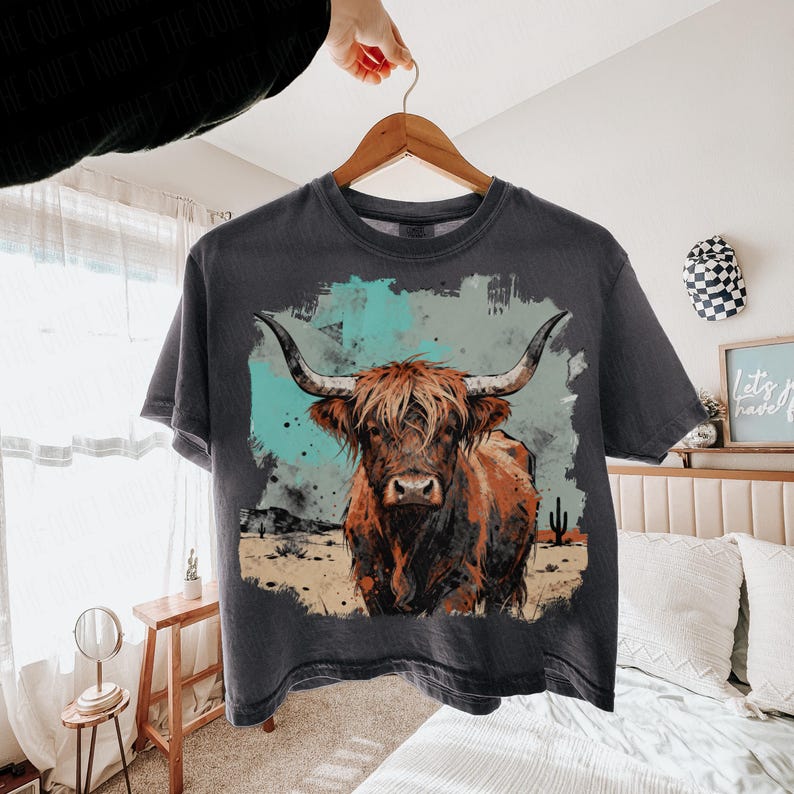 May include: Dark grey t-shirt with a watercolour image of a Highland cow. The cow is brown and tan, with long horns, set against a turquoise and tan background. The shirt is displayed on a wooden hanger.