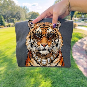 K&ouml;nnte beinhalten: Ein dunkelgraues T-Shirt mit einer detaillierten Illustration eines Tigers, der eine Sonnenbrille und eine Schmuckkette tr&auml;gt. Der Tiger hat orange und schwarze Streifen mit braun get&ouml;nten Sonnenbrillen. Die Halskette hat gro&szlig;e, klare Edelsteine.