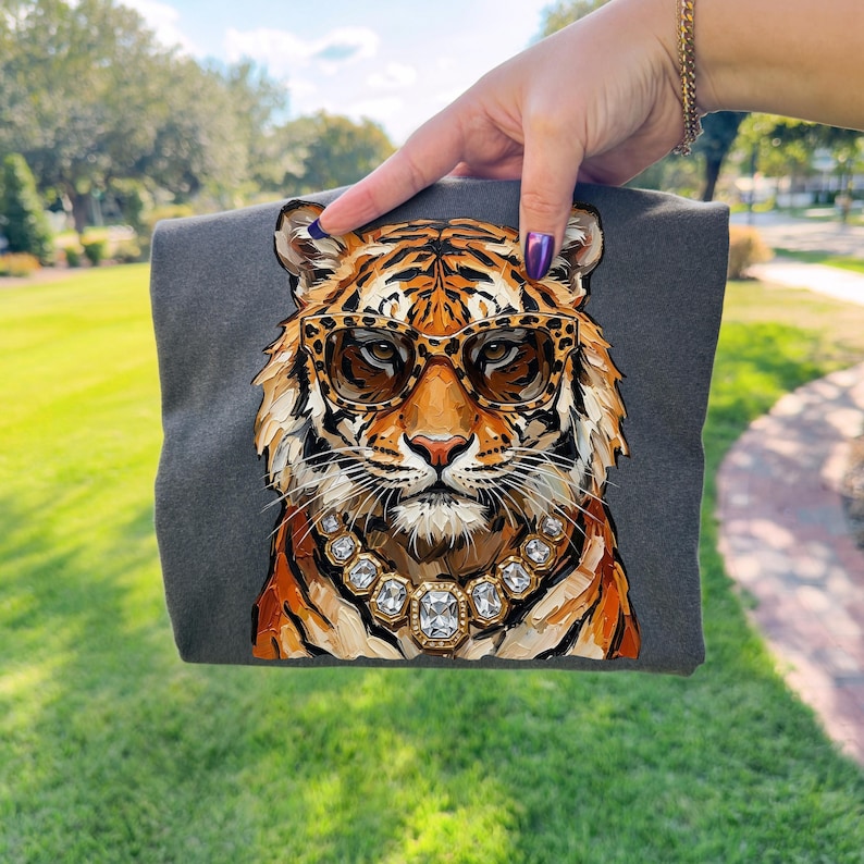 K&ouml;nnte beinhalten: Ein dunkelgraues T-Shirt mit einer detaillierten Illustration eines Tigers, der eine Sonnenbrille und eine Schmuckkette tr&auml;gt. Der Tiger hat orange und schwarze Streifen mit braun get&ouml;nten Sonnenbrillen. Die Halskette hat gro&szlig;e, klare Edelsteine.