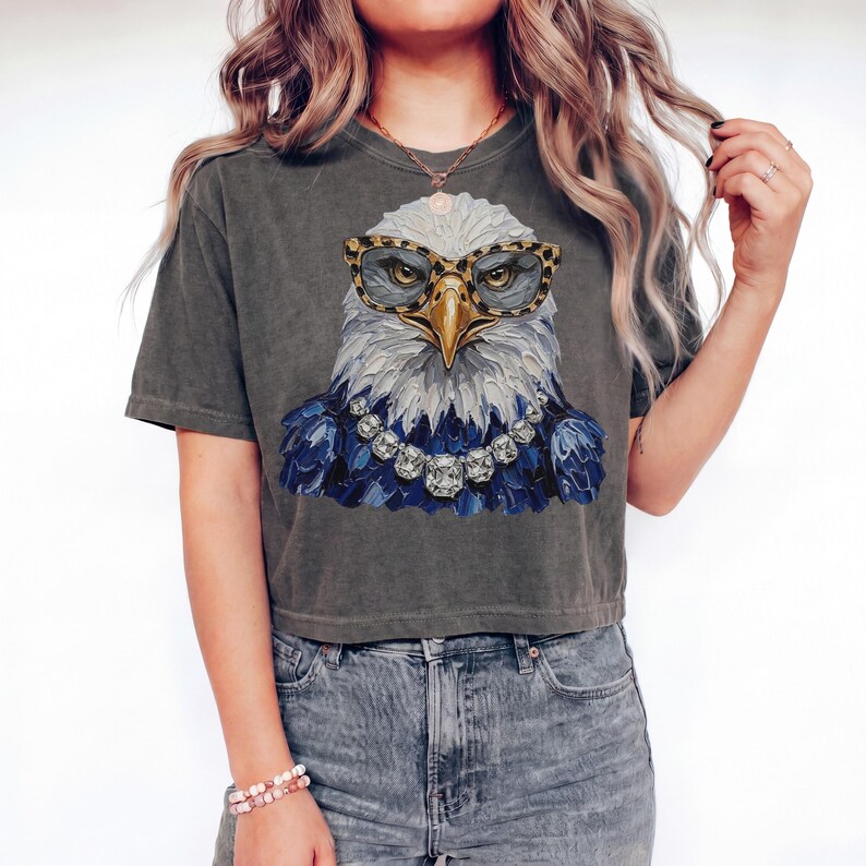 May include: A charcoal gray short-sleeve t-shirt featuring a graphic print of a bald eagle wearing leopard print glasses and a silver-toned statement necklace with large faceted crystals. The eagle has white head feathers and blue chest feathers.