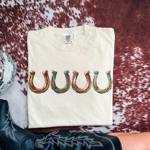 May include: Cream-colored Comfort Colors t-shirt featuring a row of five horseshoe graphics in shades of brown, blue, and orange. The shirt is laid flat on a brown and white cowhide pattern. A black cowboy boot and a disco ball are visible.