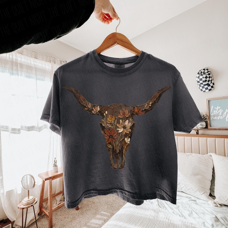 May include: Dark gray t-shirt featuring a detailed illustration of a longhorn skull adorned with colorful flowers. The artwork is centered on the shirt, creating a striking visual. The shirt is hanging on a wooden hanger.