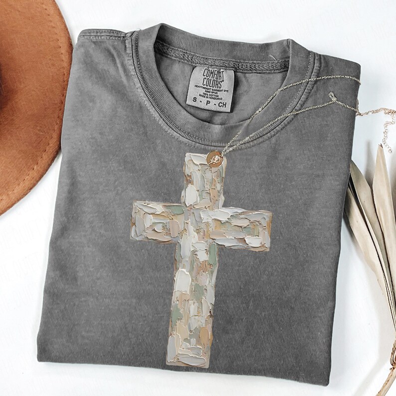 May include: A gray Comfort Colors t-shirt featuring a large, textured cross design in neutral tones. A silver necklace with a small pendant rests on the shirt. The shirt has a crew neck and short sleeves.