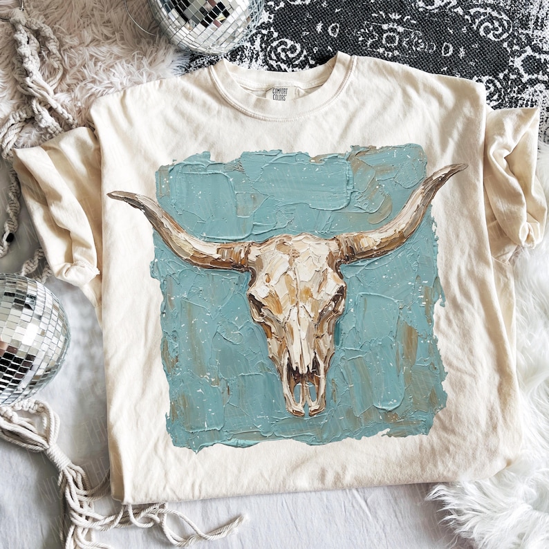 May include: Cream-coloured t-shirt with a detailed illustration of a cow skull with long horns. The skull is painted in shades of white, brown, and beige, set against a textured teal background. The shirt is a Comfort Colors brand.