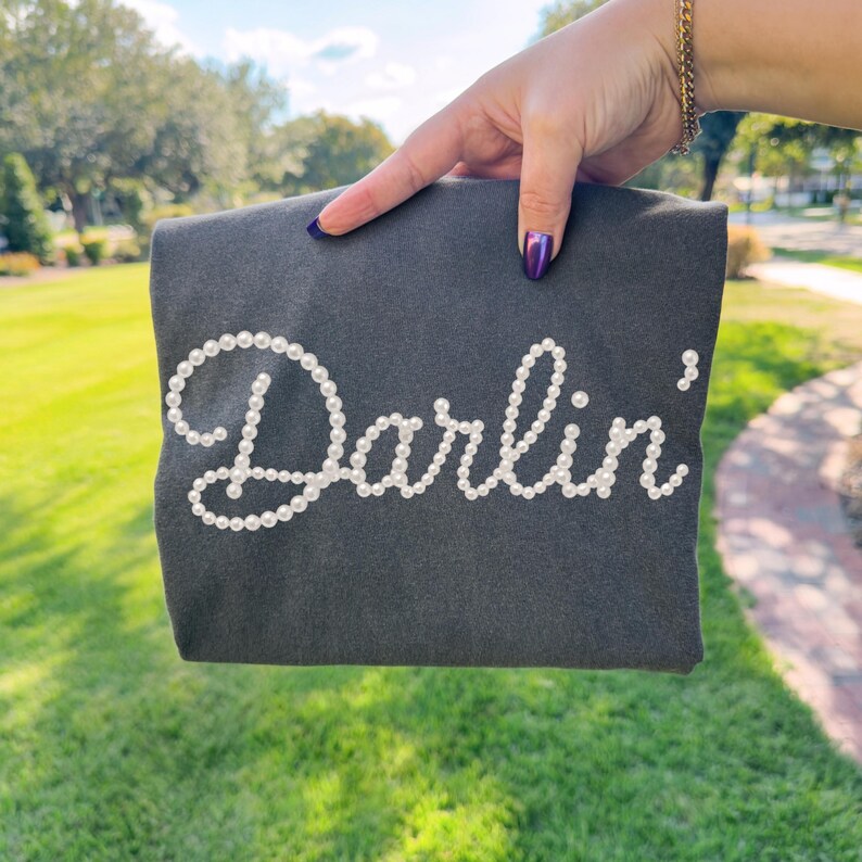 Pu&ograve; includere: Una felpa grigio scuro con la scritta "Darlin'" realizzata con perle bianche. Il capo &egrave; presentato su uno sfondo di erba verde e alberi.