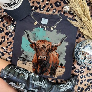 May include: A dark grey t-shirt with a Highland cow illustration, paired with a gold necklace and black cowboy boots with white embroidery. A black cap and sunglasses are also visible. The image includes a disco ball and leopard print fabric.