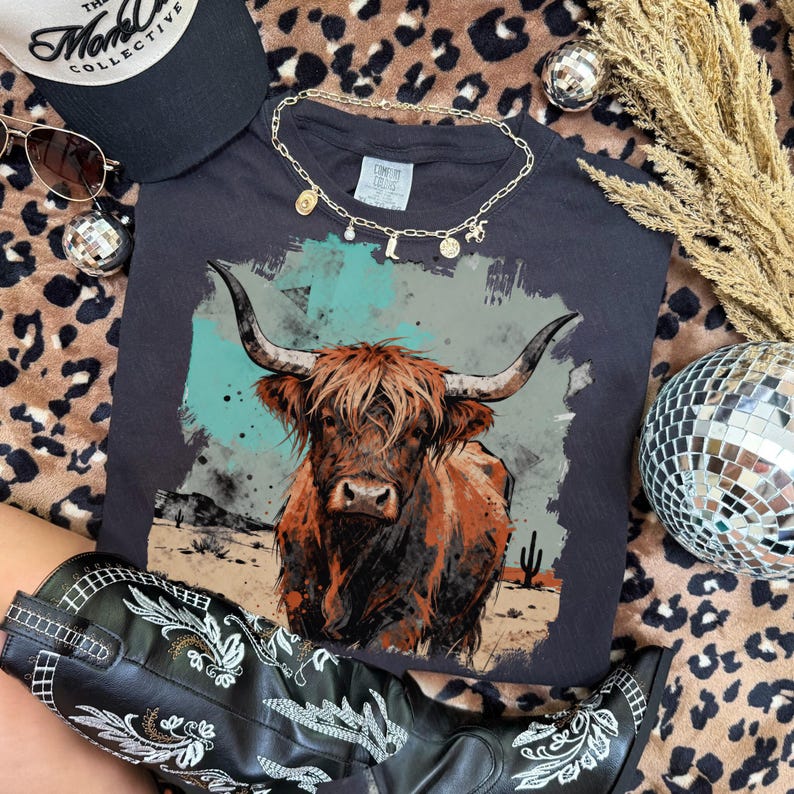 May include: A dark grey t-shirt with a Highland cow illustration, paired with a gold necklace and black cowboy boots with white embroidery. A black cap and sunglasses are also visible. The image includes a disco ball and leopard print fabric.