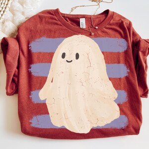 May include: A rust-colored t-shirt featuring a graphic of a cream-colored ghost with a cracked texture effect. The ghost is centered over four horizontal, distressed light purple brushstroke stripes. The shirt has a crew neckline and a tag.