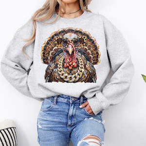 May include: A light gray crewneck sweatshirt featuring a graphic print of a turkey wearing leopard print sunglasses and a thick gold chain necklace. The turkey has detailed brown, black, and orange feathers and a prominent red wattle on its neck.