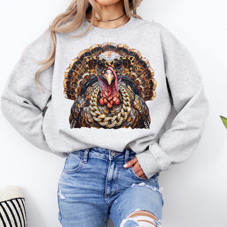 May include: A light gray crewneck sweatshirt featuring a graphic print of a turkey wearing leopard print sunglasses and a thick gold chain necklace. The turkey has detailed brown, black, and orange feathers and a prominent red wattle on its neck.