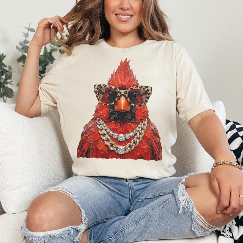 May include: Cream-colored t-shirt featuring a detailed illustration of a red cardinal bird wearing leopard print sunglasses and a diamond chain necklace. The bird has a black beak and is set against a neutral background.