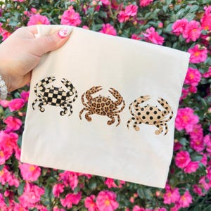 以下が含まれることがあります： 3匹のカニのイラストが描かれたクリーム色のトートバッグ。カニはそれぞれユニークな模様で、白黒のチェッカー柄、茶色のヒョウ柄、ベージュに黒い水玉模様です。バッグはピンクの花を背景に持たれています。