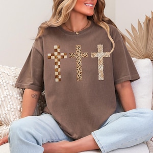 May include: Brown t-shirt featuring three crosses with unique patterns: a checkered design, a leopard print, and a dotted pattern. The shirt has a relaxed fit and is made of a soft material. The crosses are centered on the front of the shirt.