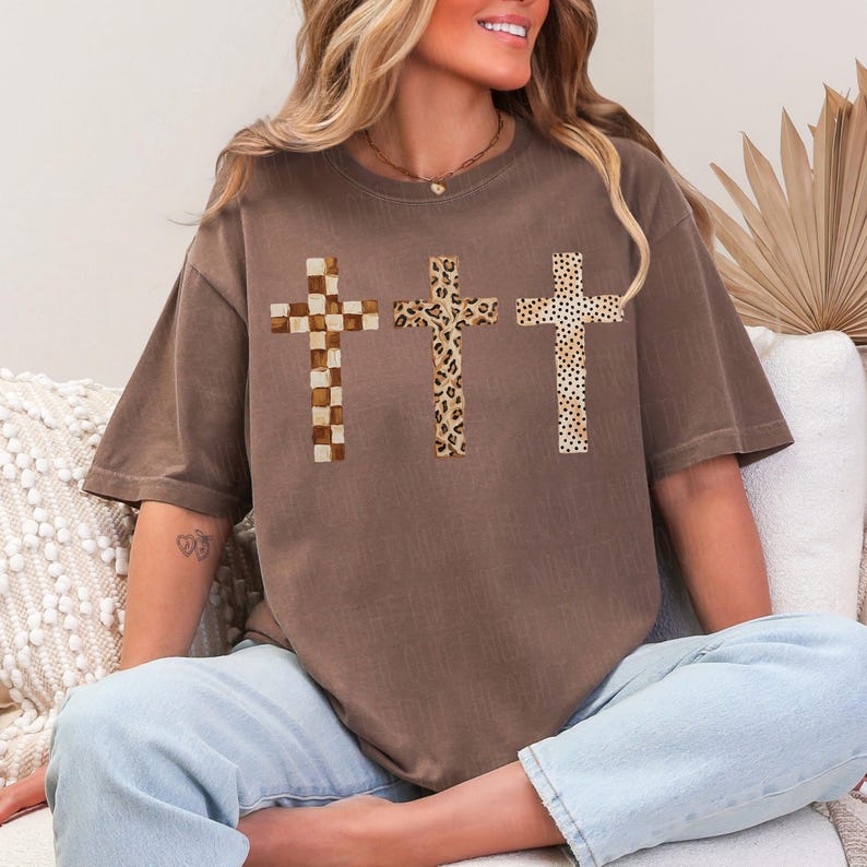 May include: Brown t-shirt featuring three crosses with unique patterns: a checkered design, a leopard print, and a dotted pattern. The shirt has a relaxed fit and is made of a soft material. The crosses are centered on the front of the shirt.