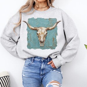 May include: A light grey sweatshirt with a detailed illustration of a cow skull and long horns. The skull is set against a textured teal background, creating a rustic and artistic design. The sweatshirt is a comfortable, casual style.