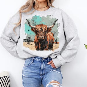 May include: A light grey sweatshirt with a watercolour-style image of a Highland cow. The cow is brown with large horns, set against a desert scene with turquoise and tan hues. The sweatshirt has long sleeves and a crew neck.
