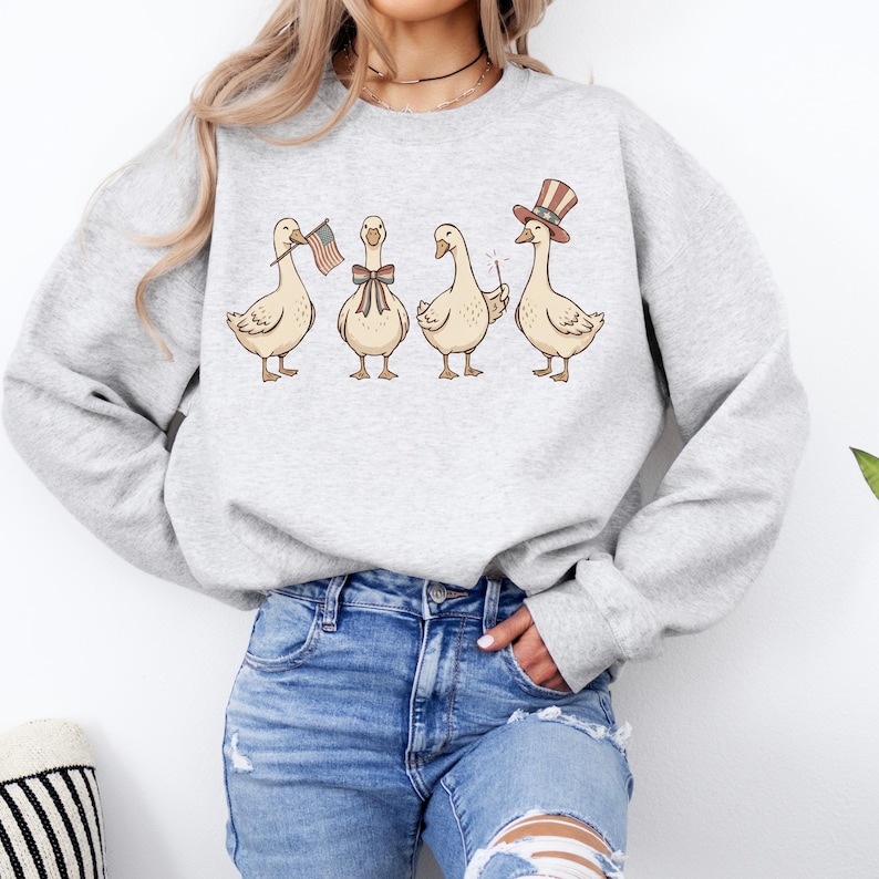 May include: A light gray sweatshirt featuring a whimsical illustration of four geese. Each goose is adorned with patriotic accessories: an American flag, a bow tie, a magic wand, and a top hat. The design is printed on a soft, comfortable fabric.