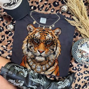 K&ouml;nnte beinhalten: Ein dunkelgraues T-Shirt mit einer detaillierten Illustration eines Tigers mit Sonnenbrille und Diamantkette. Das Bild ist mit einer schwarzen Kappe, Sonnenbrille, einer goldenen Halskette und schwarzen Cowboy-Stiefeln mit wei&szlig;er Stickerei versehen.