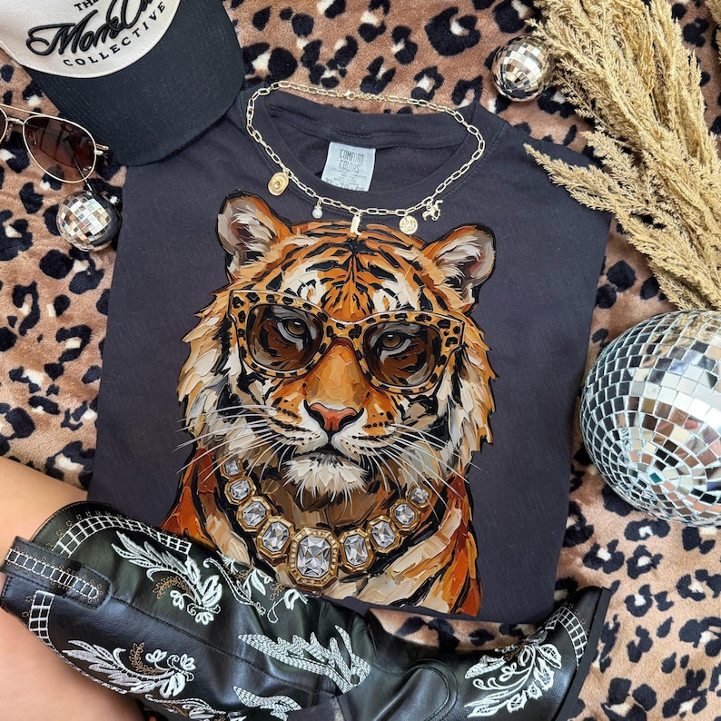 K&ouml;nnte beinhalten: Ein dunkelgraues T-Shirt mit einer detaillierten Illustration eines Tigers mit Sonnenbrille und Diamantkette. Das Bild ist mit einer schwarzen Kappe, Sonnenbrille, einer goldenen Halskette und schwarzen Cowboy-Stiefeln mit wei&szlig;er Stickerei versehen.