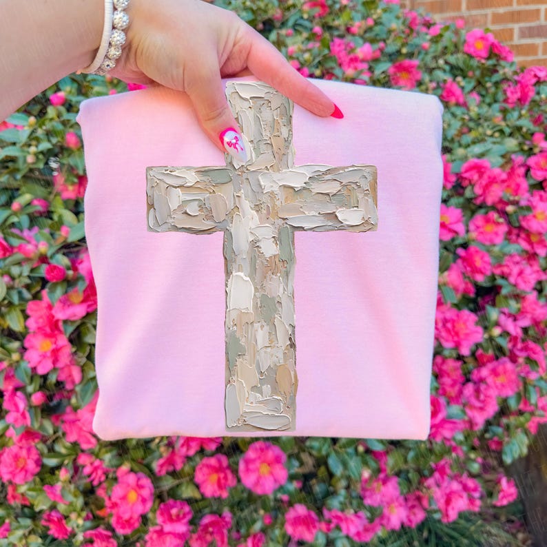 May include: A light pink sweatshirt featuring a large, textured cross design in shades of beige, gray, and white. The cross is the focal point, set against the soft pink fabric. The sweatshirt is held up by a hand.