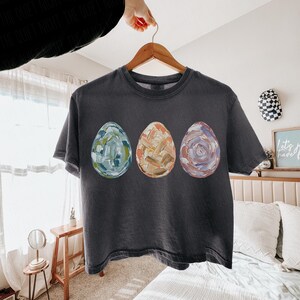 Peut inclure: T-shirt gris fonc&eacute; avec trois &oelig;ufs de P&acirc;ques peints en bleu, orange et violet. Les &oelig;ufs pr&eacute;sentent un motif textur&eacute; au pinceau. Le t-shirt est suspendu &agrave; un cintre en bois.