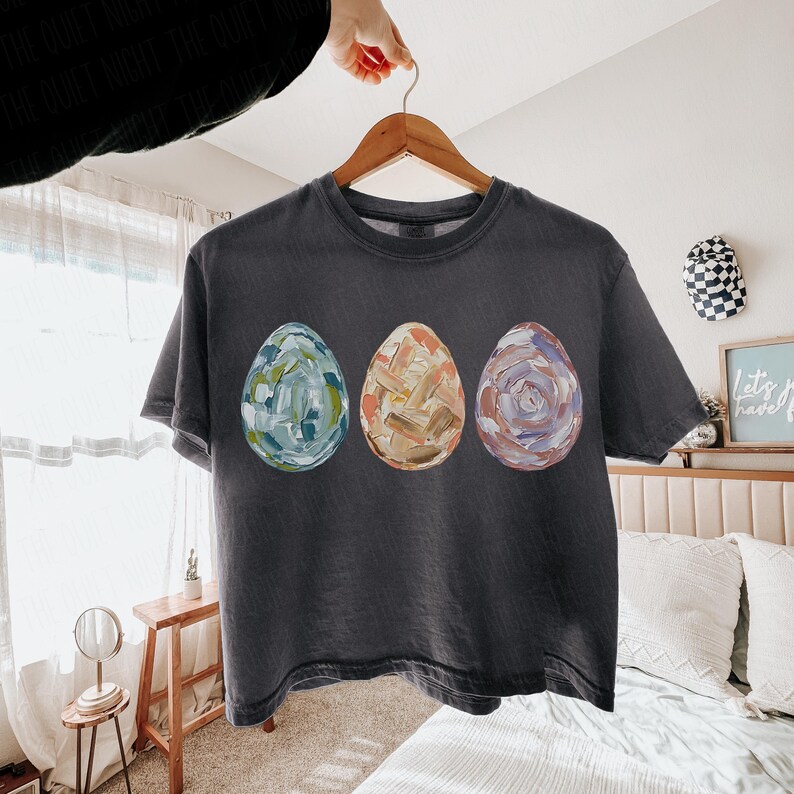 Peut inclure: T-shirt gris fonc&eacute; avec trois &oelig;ufs de P&acirc;ques peints en bleu, orange et violet. Les &oelig;ufs pr&eacute;sentent un motif textur&eacute; au pinceau. Le t-shirt est suspendu &agrave; un cintre en bois.