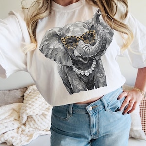 May include: A white short-sleeve t-shirt featuring a gray elephant graphic with a textured, painterly style. The elephant wears leopard-print glasses and a silver-toned necklace with large, faceted clear stones. The design is centered on the chest.