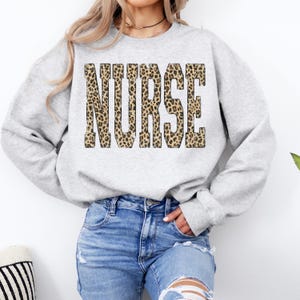 May include: A light gray sweatshirt with the word "NURSE" in large, leopard print letters across the chest. The sweatshirt has long sleeves and a crew neck. The model is wearing blue jeans.