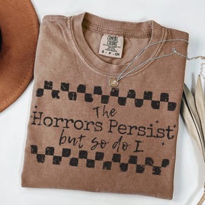 May include: A brown Comfort Colors t-shirt with the text "The Horrors Persist but so do I" in black. The shirt has a checkerboard pattern above and below the text. A silver necklace with a pendant is draped across the shirt.
