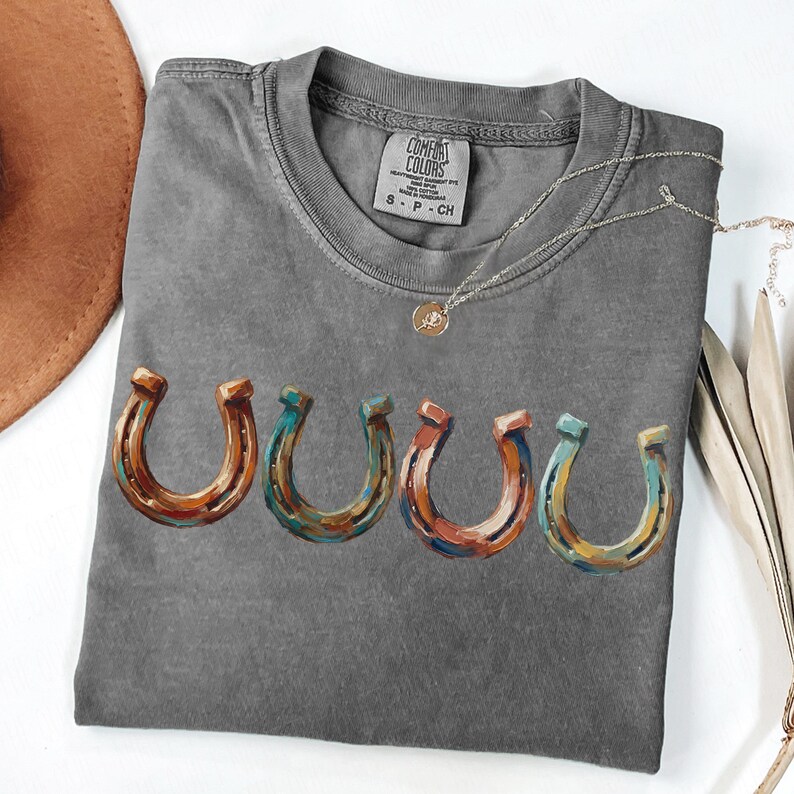 May include: A gray Comfort Colors t-shirt featuring a row of five colorful horseshoe graphics. A silver necklace with a small pendant is draped across the shirt. The shirt is folded, and a brown hat is visible in the upper left corner.