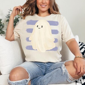 May include: A cream-colored short-sleeve t-shirt featuring a central graphic of a pale yellow ghost with a simple smiling face. The ghost is positioned over a background of four horizontal, textured, periwinkle blue brushstroke stripes.