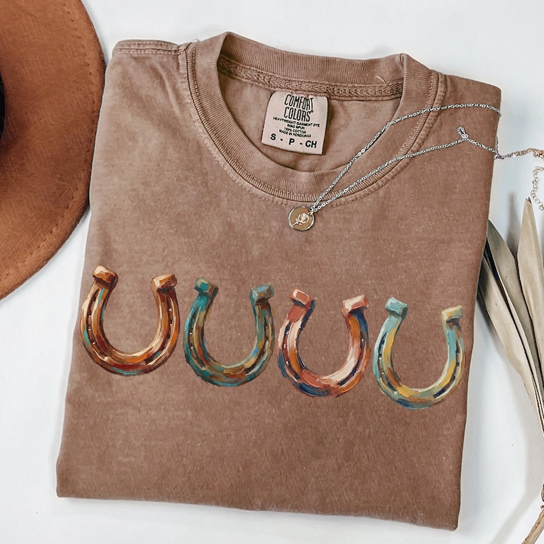 May include: Brown Comfort Colors t-shirt with a crew neck and the word "UUUU" printed in colorful horseshoe designs. A silver necklace with a pendant is draped across the shirt. The shirt is size S-P-CH.
