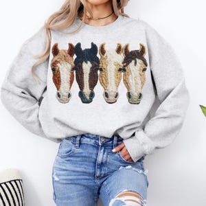 Peut inclure: Un sweat-shirt gris clair orn&eacute; de quatre t&ecirc;tes de chevaux brod&eacute;es dans des tons de marron, noir et cr&egrave;me. Les t&ecirc;tes de chevaux sont dispos&eacute;es horizontalement. Le sweat-shirt est port&eacute; avec un jean bleu.