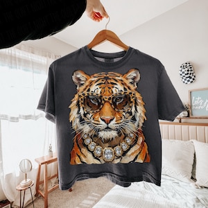 K&ouml;nnte beinhalten: Dunkelgraues T-Shirt mit einem Tiger, der eine Leopardenbrille und eine Diamantkette tr&auml;gt. Der Tiger hat orange-schwarzes Fell. Das T-Shirt h&auml;ngt an einem Holzb&uuml;gel.