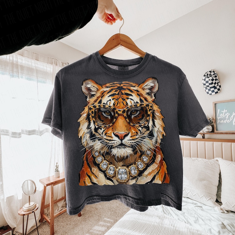 K&ouml;nnte beinhalten: Dunkelgraues T-Shirt mit einem Tiger, der eine Leopardenbrille und eine Diamantkette tr&auml;gt. Der Tiger hat orange-schwarzes Fell. Das T-Shirt h&auml;ngt an einem Holzb&uuml;gel.