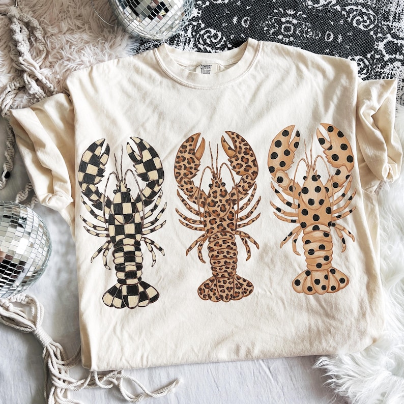 May include: Cream-colored t-shirt featuring three lobster illustrations. The lobsters have unique patterns: checkered, leopard print, and dotted. The shirt has a crew neck and short sleeves. The brand name "Comfort Colors" is visible on the tag.