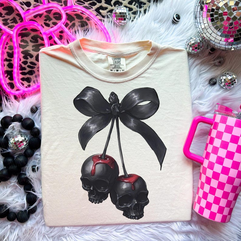 May include: A cream-coloured t-shirt with a black bow and two cherries with skull faces and red drips. The shirt has a crew neck and short sleeves. The brand label is visible at the neckline.