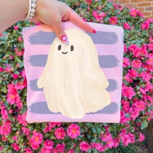 May include: A light pink sweatshirt featuring a central graphic of a cream-colored ghost with a simple smiling face, set against a background of horizontal, distressed-style purple brushstroke stripes across the fabric.