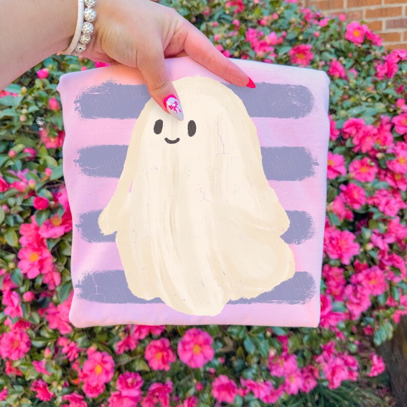 May include: A light pink sweatshirt featuring a central graphic of a cream-colored ghost with a simple smiling face, set against a background of horizontal, distressed-style purple brushstroke stripes across the fabric.