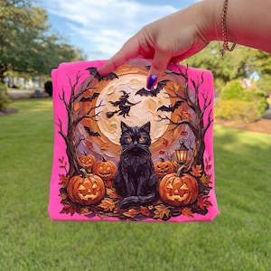 May include: A bright neon pink sweatshirt featuring a detailed graphic print of a black cat sitting in front of a large full moon, surrounded by carved jack-o'-lanterns, bare trees, flying bats, and a silhouette of a witch riding a broomstick.