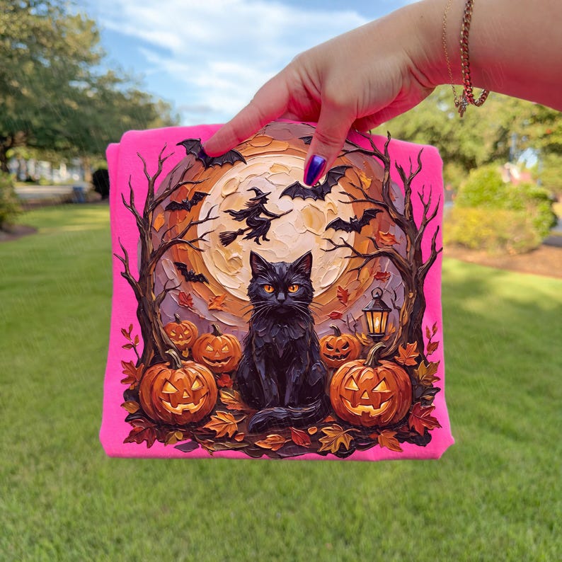 May include: A bright neon pink sweatshirt featuring a detailed graphic print of a black cat sitting in front of a large full moon, surrounded by carved jack-o'-lanterns, bare trees, flying bats, and a silhouette of a witch riding a broomstick.