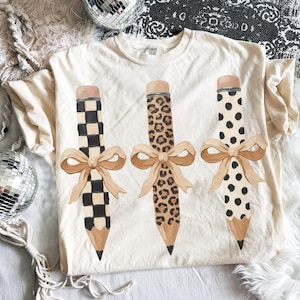 以下が含まれることがあります： クリーム色のTシャツ。3つの鉛筆のデザインとリボンが描かれています。1つの鉛筆は白と黒の市松模様、もう1つはヒョウ柄、3つ目は黒い水玉模様です。各鉛筆にはベージュのリボンが付いています。