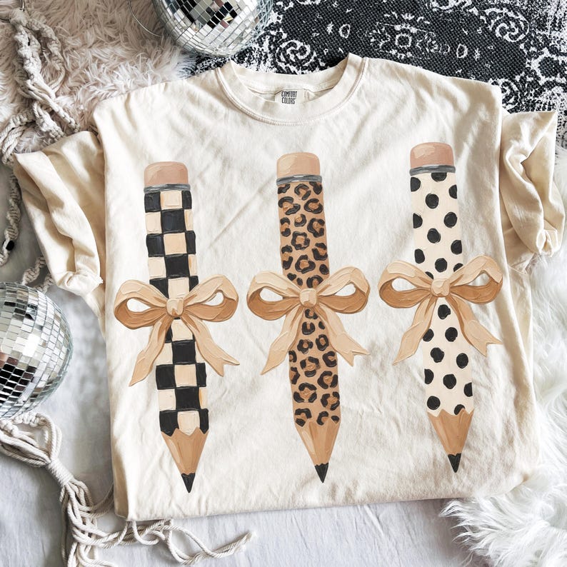 以下が含まれることがあります： クリーム色のTシャツ。3つの鉛筆のデザインとリボンが描かれています。1つの鉛筆は白と黒の市松模様、もう1つはヒョウ柄、3つ目は黒い水玉模様です。各鉛筆にはベージュのリボンが付いています。