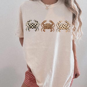以下が含まれることがあります： 3匹のカニのイラストが描かれたクリーム色のTシャツ。それぞれ市松模様、レオパード柄、水玉模様と異なるパターン。ゆったりとしたフィット感で、半袖です。