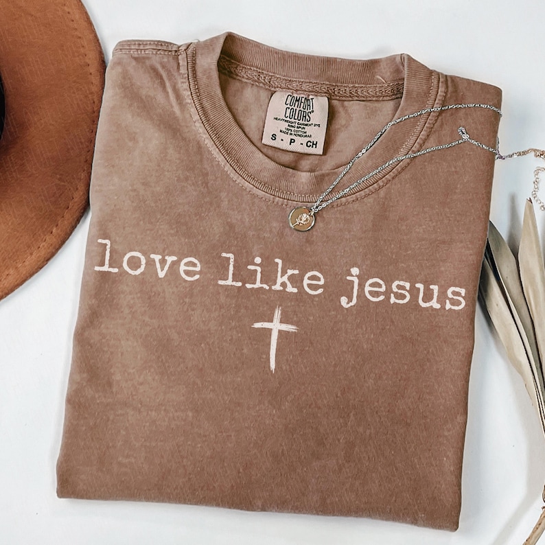 May include: A brown Comfort Colors t-shirt with the words "love like jesus" and a cross printed in white. A silver necklace with a pendant is draped across the shirt. A brown hat is visible in the upper left corner.