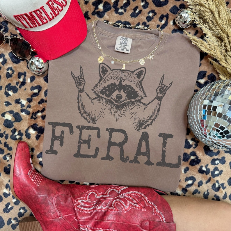 May include: A brown t-shirt with a raccoon graphic giving the rock on hand gesture and the word "FERAL" printed below. The image also includes a red baseball cap, sunglasses, a gold necklace, a disco ball, and red cowboy boots.